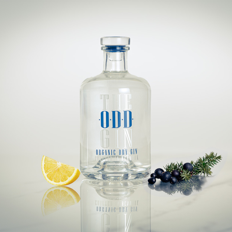 The ODD Gin med en ginflaske – Organic Dry - placeret ved siden af enebærgren og citronskive i et lyst marmor lignende setup