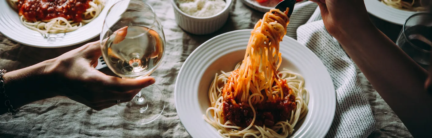 Person spiser spaghetti med kødsovs fra en tallerken, mens der holdes et glas hvidvin ved siden af, som stemningsbillede til vinvalg til pastaretter