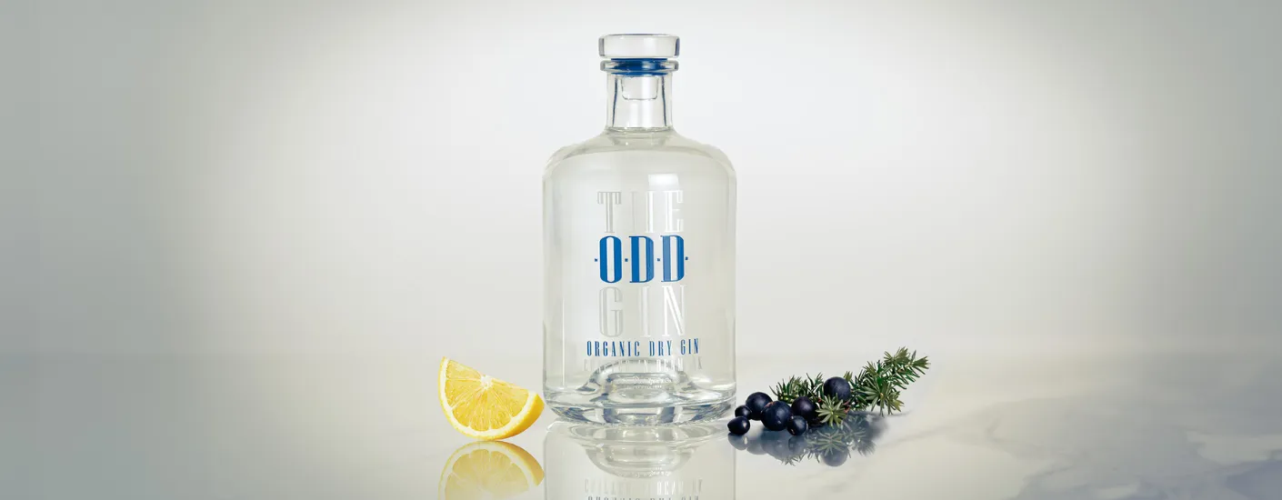 The ODD Gin med en ginflaske – Organic Dry - placeret ved siden af enebærgren og citronskive i et lyst marmor lignende setup